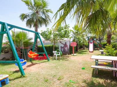 Kinderspielplatz mit Schaukel, Rutsche, Sitzbänken und Palmen im Garten unter blauem Himmel.