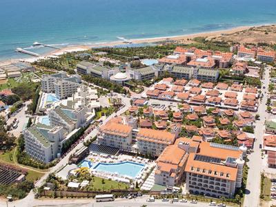 Blick auf eine Küstenstadt mit Hotels, roten Dächern, Pool und Strand am blauen Meer.