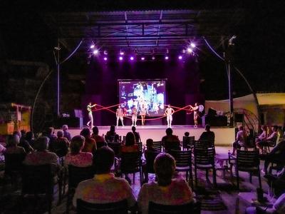 Theaterbühne mit Lichtern und Darstellern, Publikum sitzt im Dunkeln, abends, lebhafte Atmosphäre.