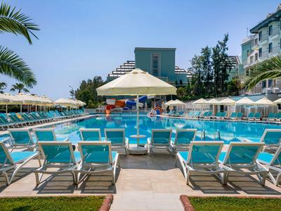 Duży obszar basenu z niebieskimi leżakami, parasolami i nowoczesnymi budynkami w tle.