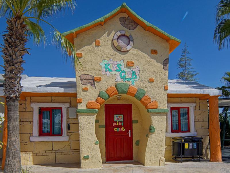 Edificio colorato del club per bambini con porta rossa, orologio e palme sotto un cielo limpido.