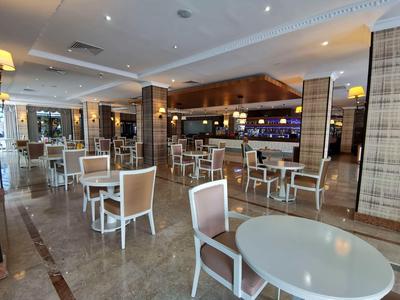 Area pranzo moderna con sedie e tavoli bianchi, pavimento in marmo chiaro e bar sullo sfondo.