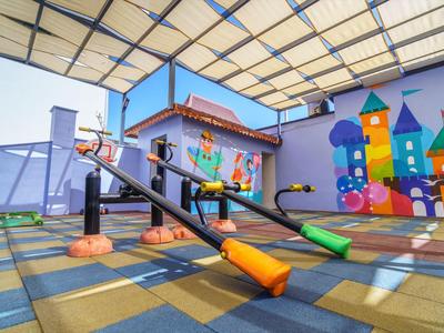 Area giochi colorata con altalene a bilico sotto una zona coperta accanto a un murale colorato.