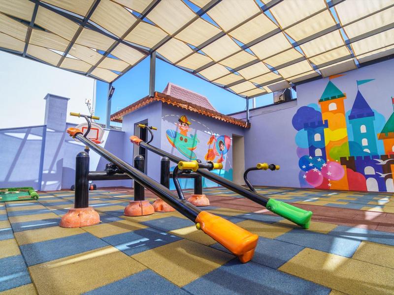 Area giochi colorata con altalene a bilico sotto una zona coperta accanto a un murale colorato.