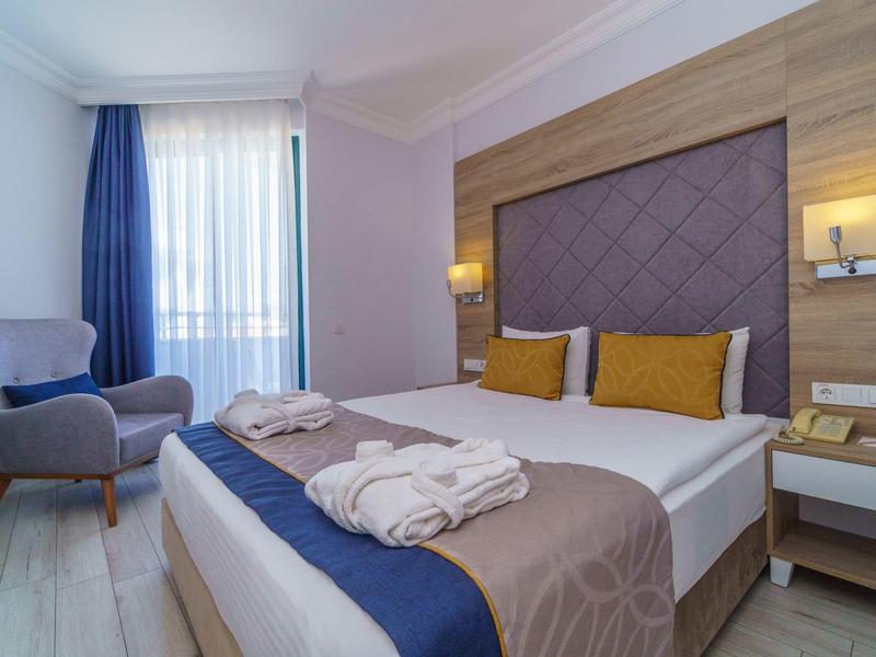 Camera d'hotel moderna con letto matrimoniale, due cuscini, accappatoi e poltrona accanto a una finestra.