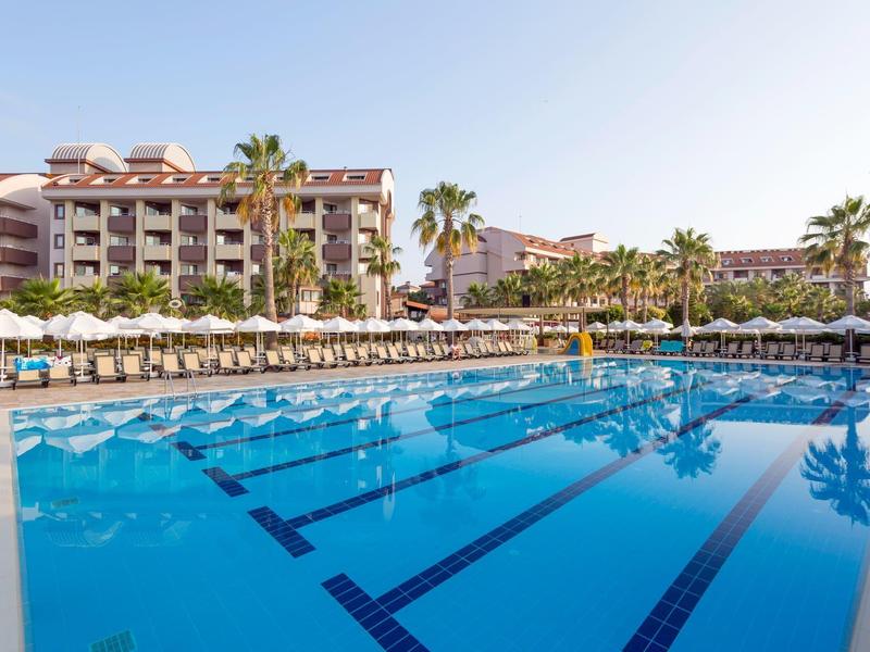 Large hotel with outdoor pool, palm trees, and numerous lounge chairs under clear sky.