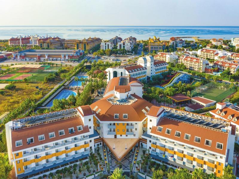 Aerial view of a hotel with red roofs, surrounded by greenery and sea in the background.