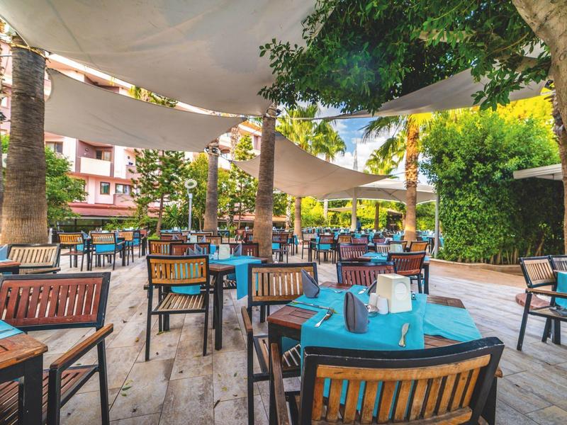 Outdoor restaurant with blue tablecloths, wooden tables and chairs under white sun sails and palm trees.