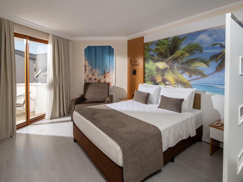 Bright hotel room with large bed, armchair, balcony, and beach wall art.