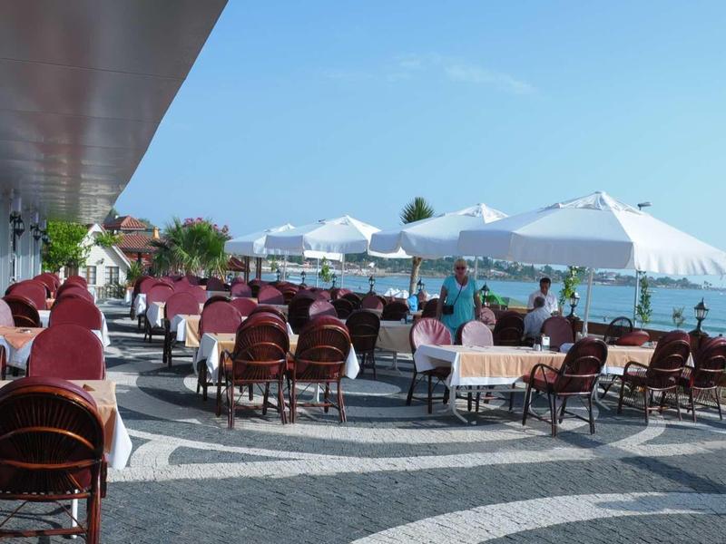 Outdoor seaside restaurant with tables, chairs, and white umbrellas along a promenade.