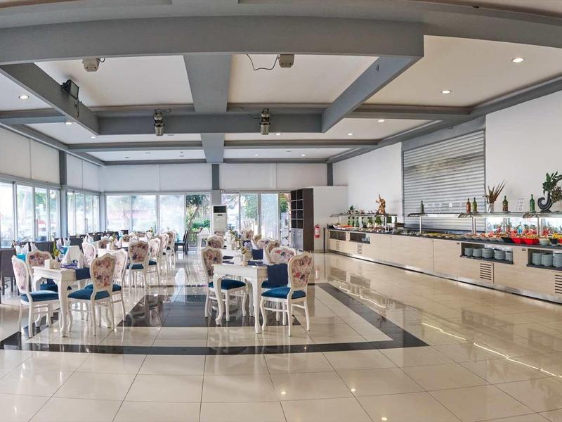Spacious hotel dining area with large windows, white chairs, and buffet counter under bright lighting.