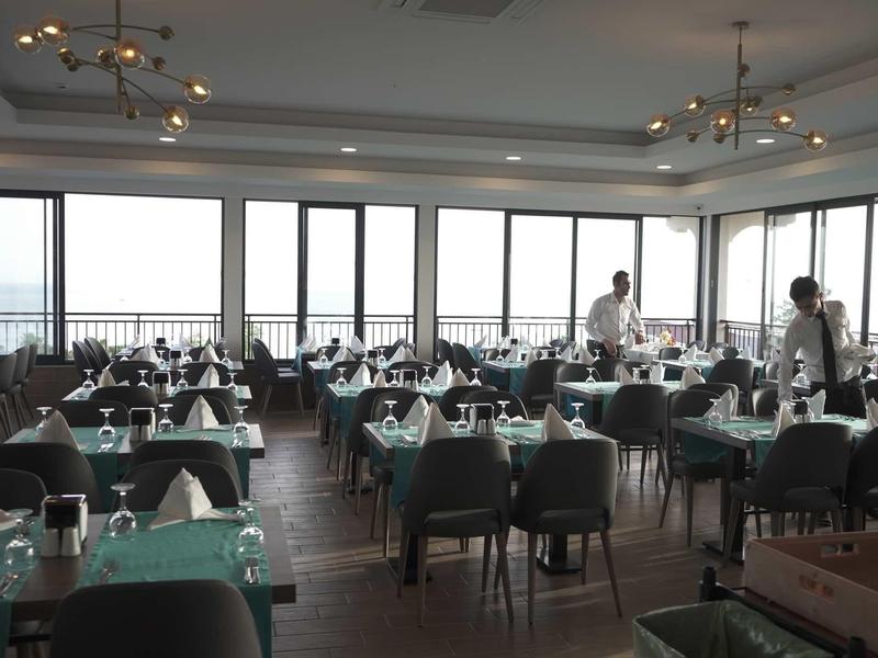 Ristorante spazioso con grandi finestre che si affacciano sull'acqua, tavoli apparecchiati e personale presente.