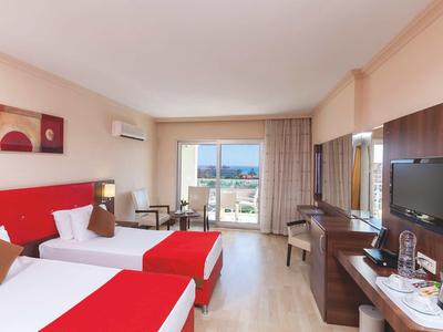 Camera d'hotel con due letti singoli, scrivania, TV e balcone con vista sul mare.