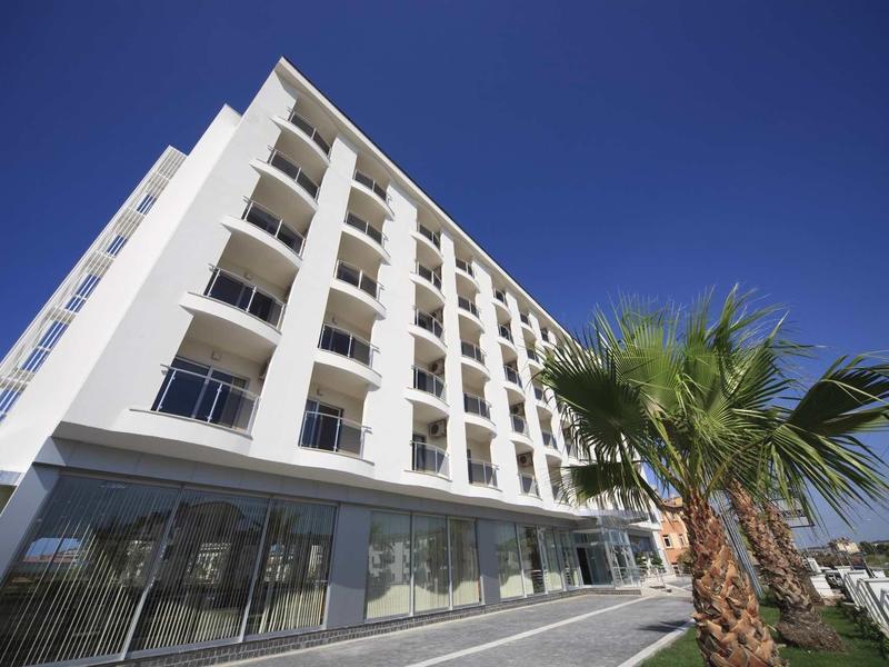 Edificio alberghiero bianco moderno con balconi sotto un cielo azzurro limpido e palme in primo piano.