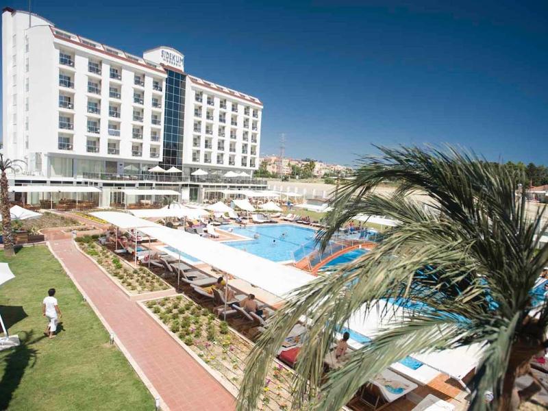 Edificio dell'hotel con piscina all'aperto, lettini e palme in una giornata di sole.