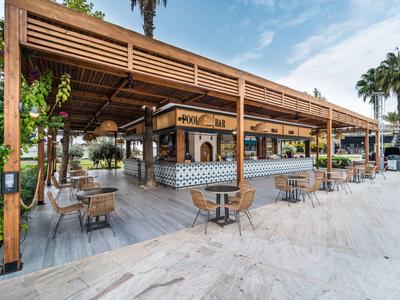 Open beach restaurant with wooden roof, tables, and chairs on sand under blue sky.