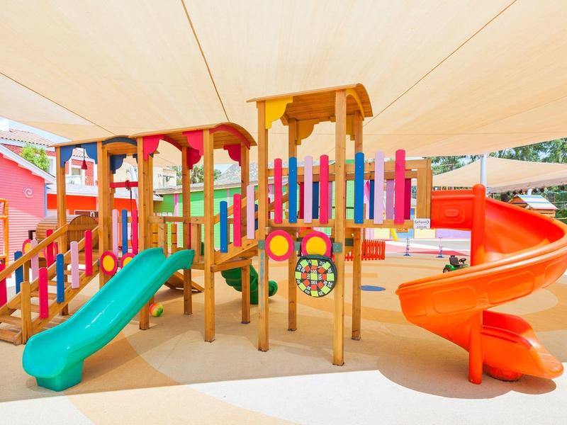 Colorful children's playground with climbing frames and slides under a sunshade on hotel grounds.