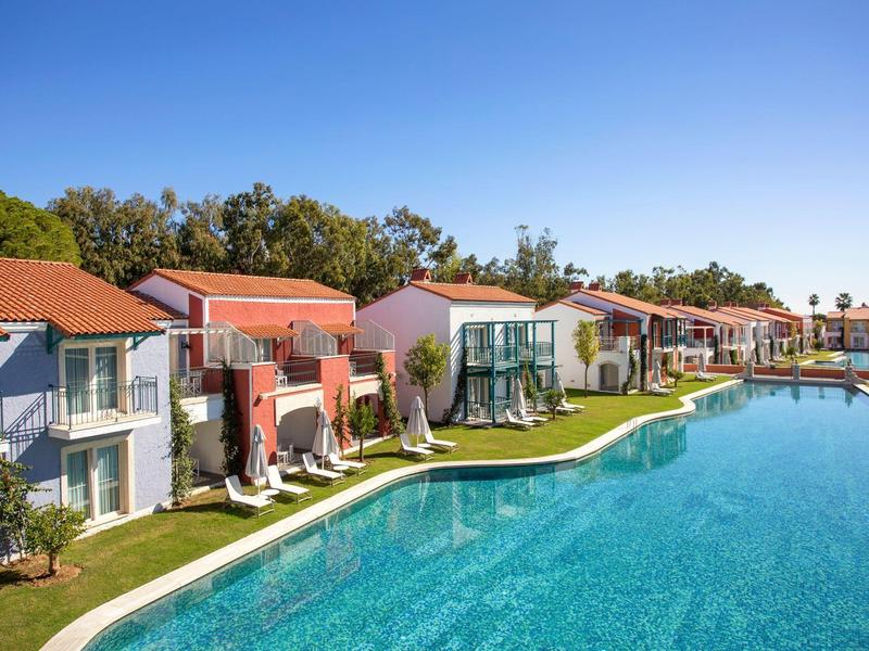 Modern villas with red roofs line a clear blue pool under a sunny sky.