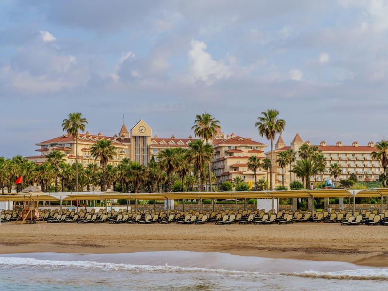 View of a hotel with palm trees and sandy beach in the foreground under blue sky.