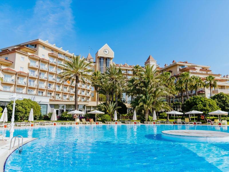 Large hotel pool with clear blue water and lounge chairs in front of a multi-story hotel building.