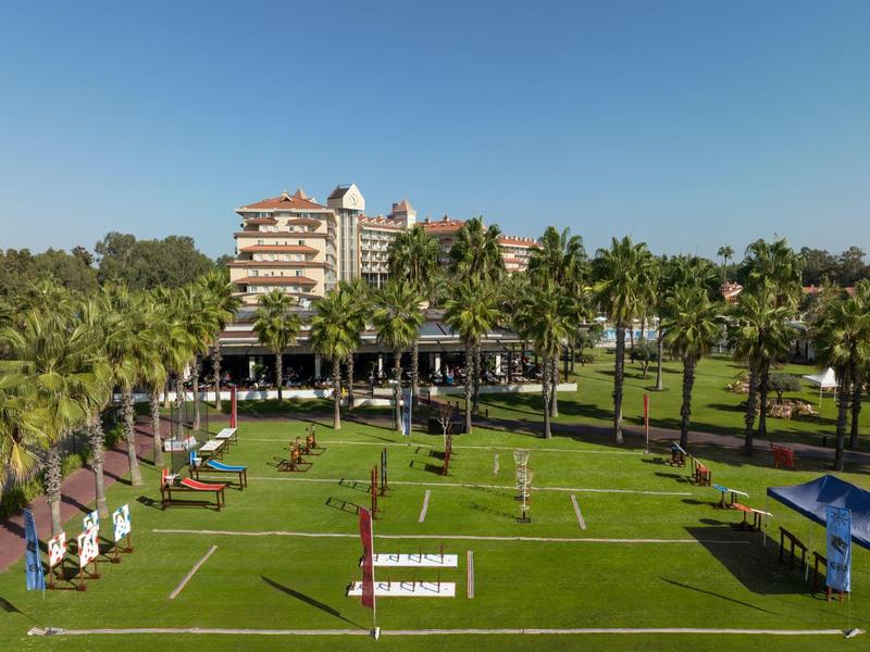Large hotel garden with volleyball court, sun loungers, and palm trees under a clear sky