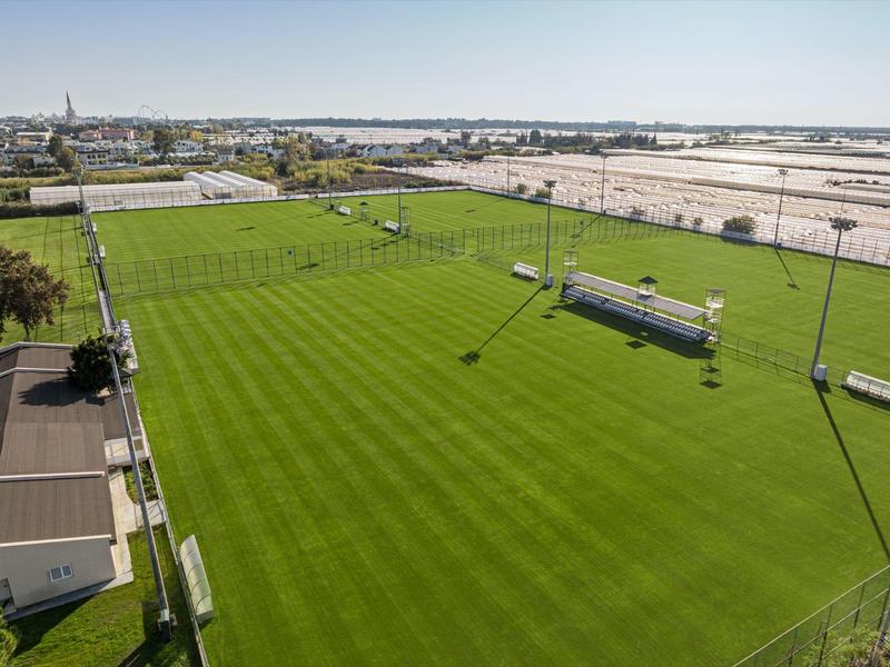 Large, well-maintained green sports field with stands and floodlights in a sunny setting
