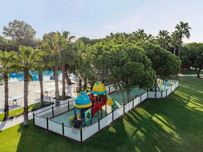 Colorful children's playground with slide in a hotel garden, surrounded by palm trees and greenery.