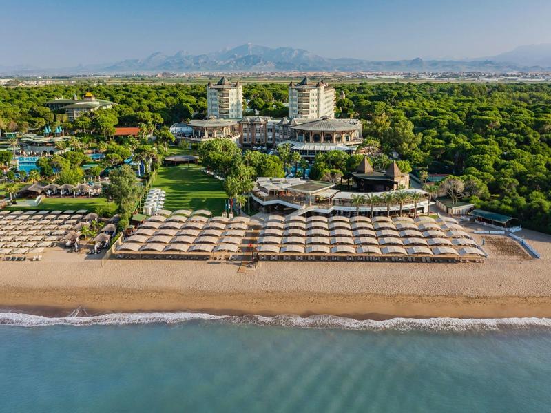 Large hotel with pool and sun umbrellas on sandy beach, surrounded by lush greenery.