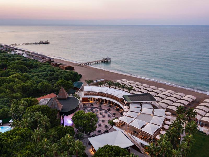 Aerial view of a beach hotel with umbrellas, pool, and forested area at sunset.