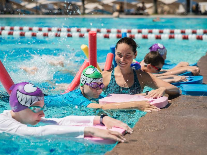 Children learning to swim using kickboards in pool under supervision.