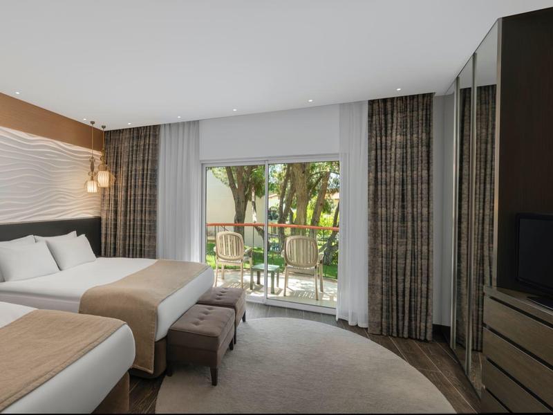 Modern hotel room with double bed, armchairs, TV, and balcony overlooking garden.