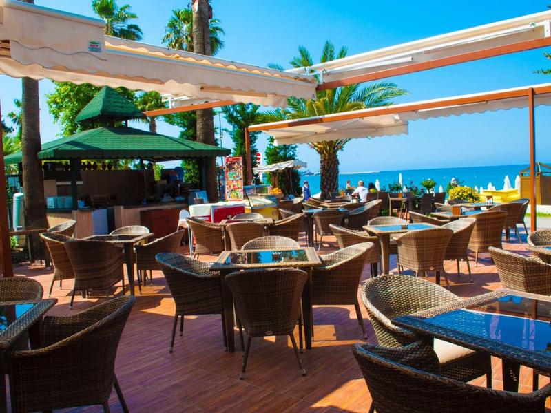 Freiluftrestaurant mit Holzterrasse, Rattanmöbeln, Sonnensegeln und Meerblick im Hintergrund.
