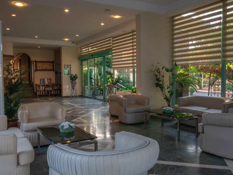 Moderner, hell beleuchteter Wohnraum mit hellen Sofas, runden Tischen und großen Fenstern mit Jalousien.