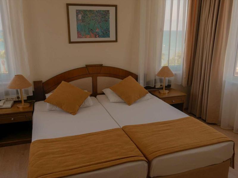 Ein helles Schlafzimmer mit zwei Einzelbetten, braunen Kissen, Vorhängen und zwei Nachttischen mit Lampen.