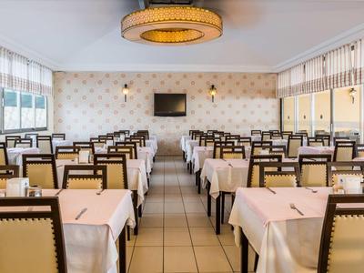 Großer heller Speisesaal mit rechteckigen Tischen, beigen Tischdecken und Holzstühlen mit Polster.