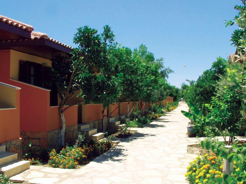 Sunny paved pathway between red houses and green trees in a holiday resort.