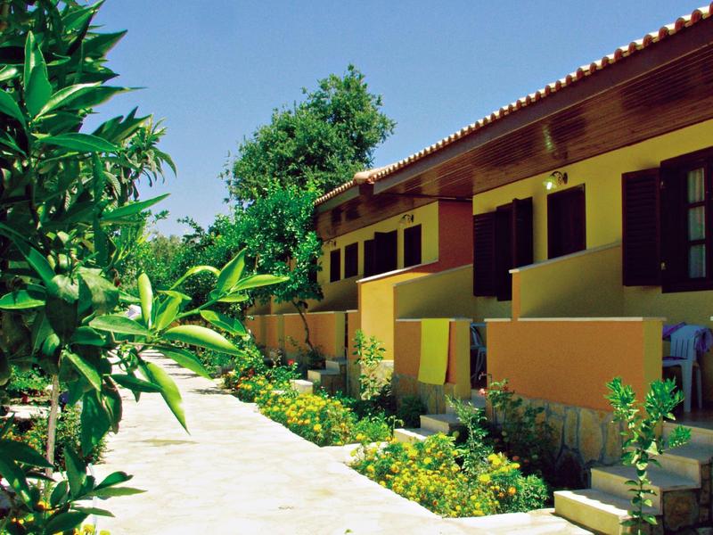 Modern hotel walkway with planted garden and yellow building with red roofs under clear sky.