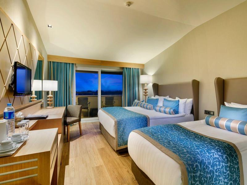 Modern hotel room with two beds, desk, and city view at dusk.