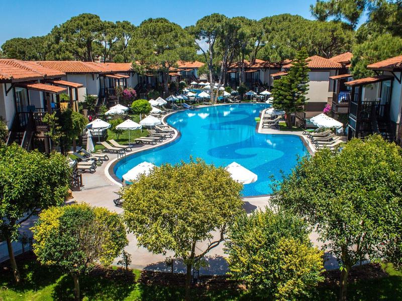 Hotel complex with curved pool, surrounded by plants and double bungalows with red roofs.