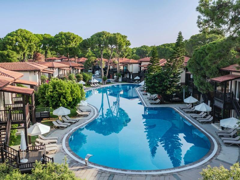 Hotel complex with large pool, sun loungers, and green garden under clear sky.