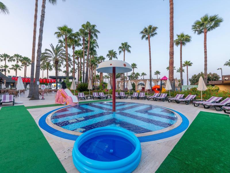 Area piscina dell'hotel con lettini e palme sotto un cielo limpido.