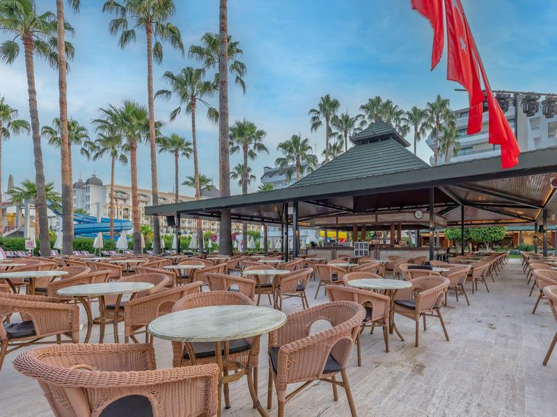 Ampia terrazza con più tavoli e sedie, circondata da palme e cielo limpido.
