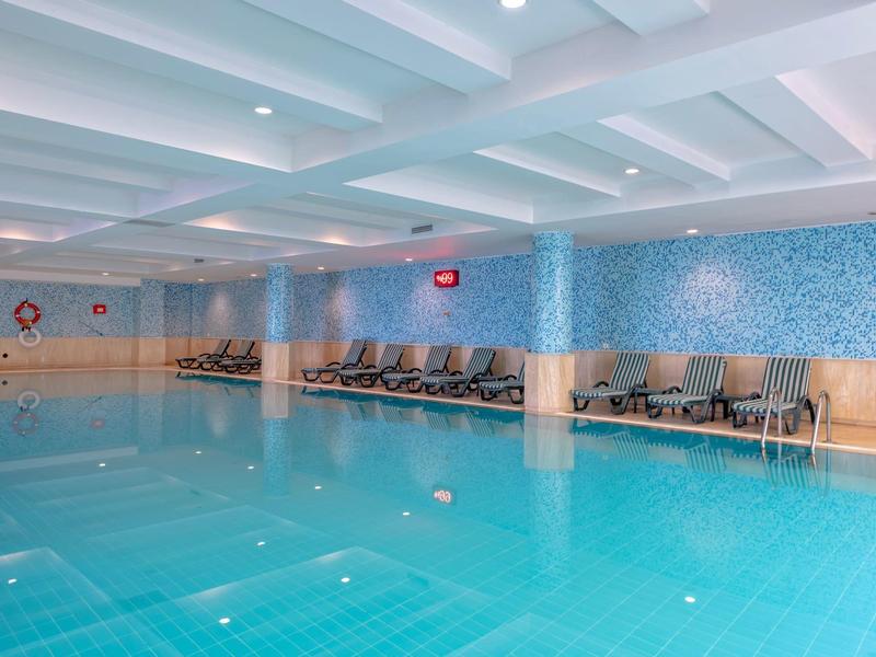 Piscina interna con piastrelle blu, lettini e luci a soffitto in un hotel.
