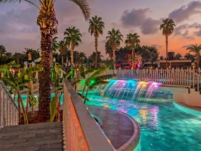 Bunte Wasserfälle fließen in einen Pool, umgeben von Palmen und einem Abendhimmel mit Wolken.