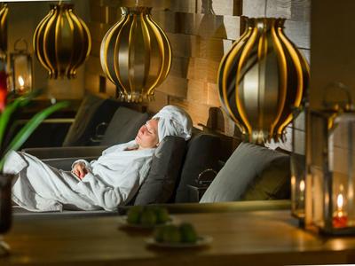 Person relaxes in white bathrobe on lounge chair in cozy indoor setting with golden lamps.
