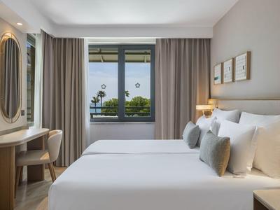 Habitación de hotel luminosa con cama doble, ventana con vista a palmeras y mobiliario moderno.