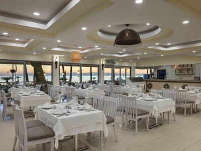 Restaurante luminoso con mesas y sillas blancas, vista al mar a través de grandes ventanas.