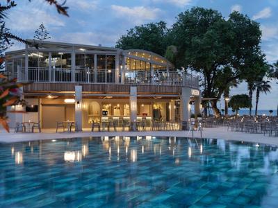 Hotel de diseño moderno iluminado por la noche, gran piscina en primer plano, palmeras al fondo.