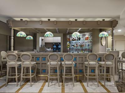 Área de bar moderna en hotel con nueve taburetes blancos y estantería retroiluminada para bebidas.