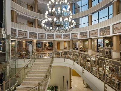 Amplio vestíbulo de hotel con escaleras, candelabro y barandales de vidrio.